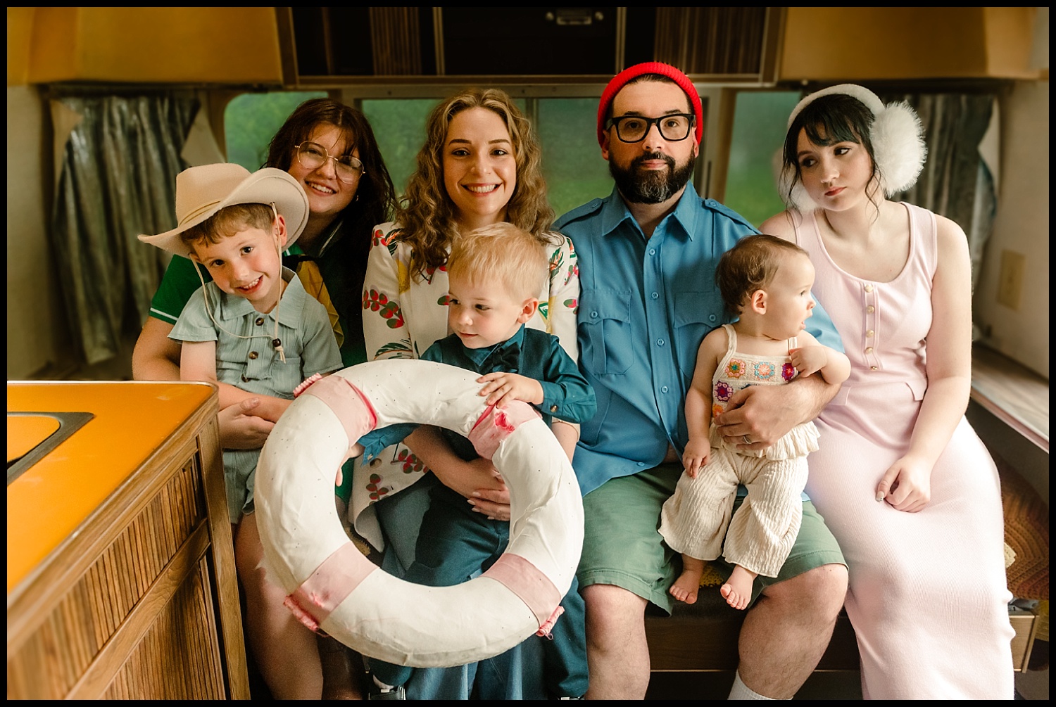 Seven people, including three children and four adults, sit closely together on a bench inside a vintage camper. Styled in colorful, Wes Anderson-inspired outfits, this Nashville group creates a quirky family portrait as one child holds a life preserver.