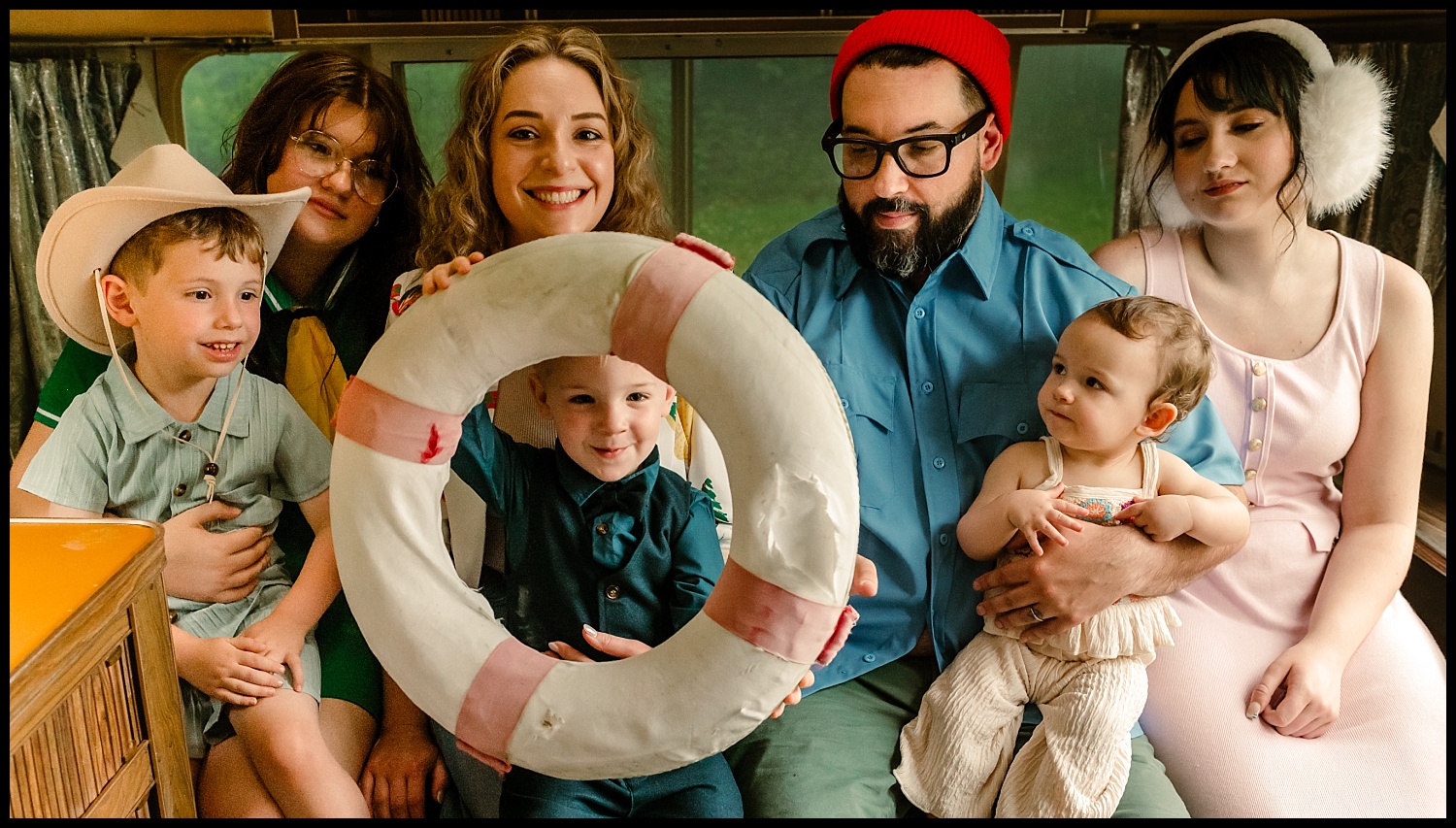 A group of six people, including two children, sit closely together indoors in true Wes Anderson style. One child holds a large, white life preserver with red stripes as the others smile for their Nashville-inspired family portrait.