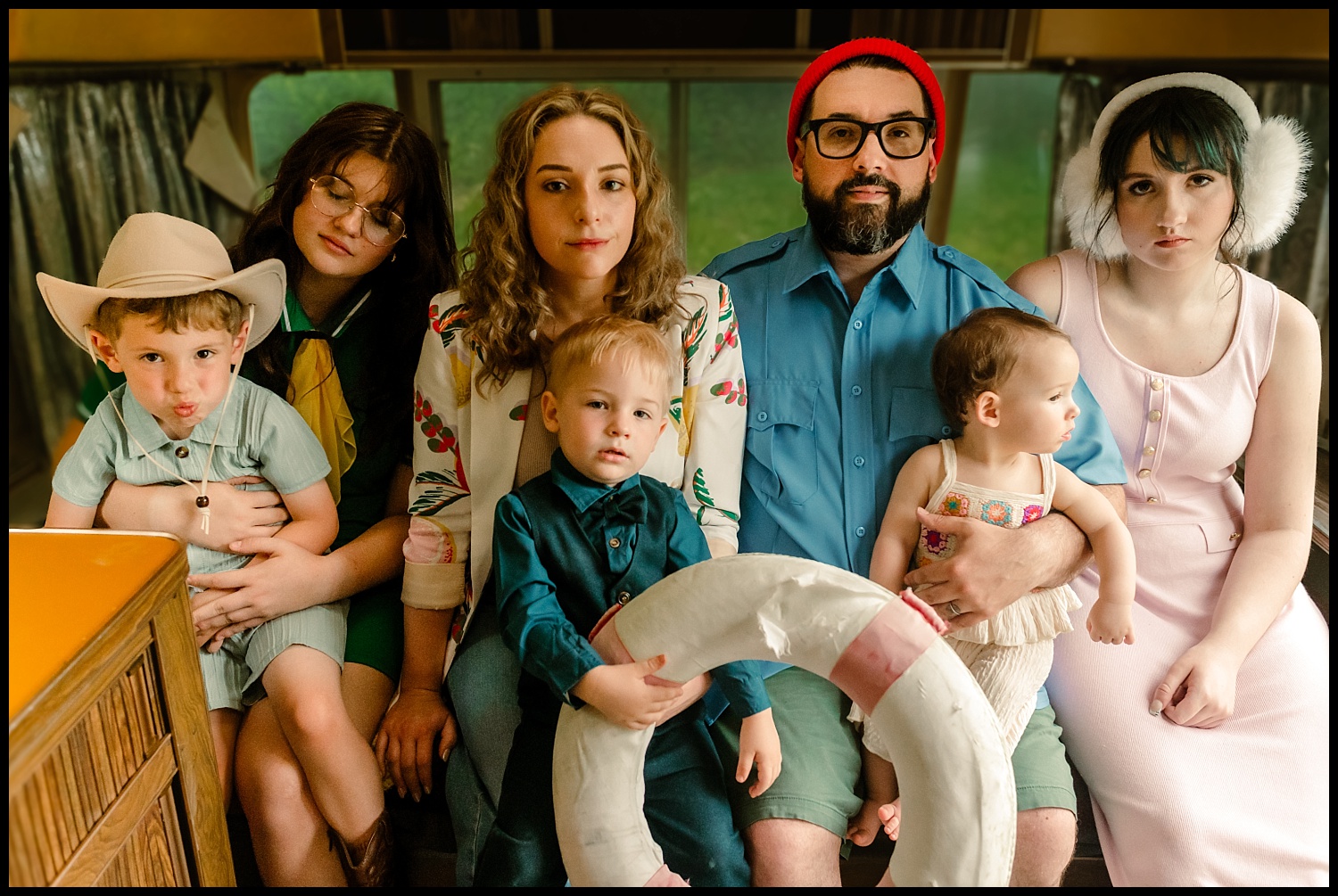 A group of six people and one baby sit close together indoors, dressed in colorful, vintage outfits reminiscent of Wes Anderson family portraits. Accessories like a cowboy hat and earmuffs add quirky flair to this serious yet stylish scene.
