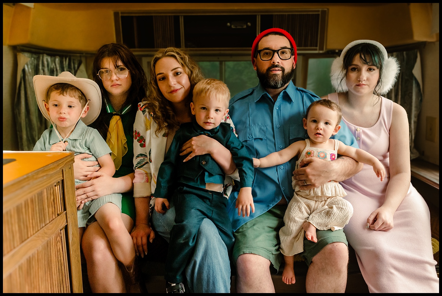 Six people, including three children and three adults, sit closely on a bench inside a retro RV in Nashville, all looking at the camera with neutral expressions—evoking the quirky style of Wes Anderson family portraits with their colorful, eclectic outfits.