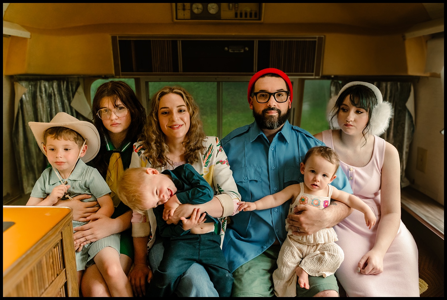 A family of six sits closely together in a retro RV, captured in true Wes Anderson style. The parents are in the middle with a crying toddler and baby, while three children sit beside them, each with unique expressions and outfits.