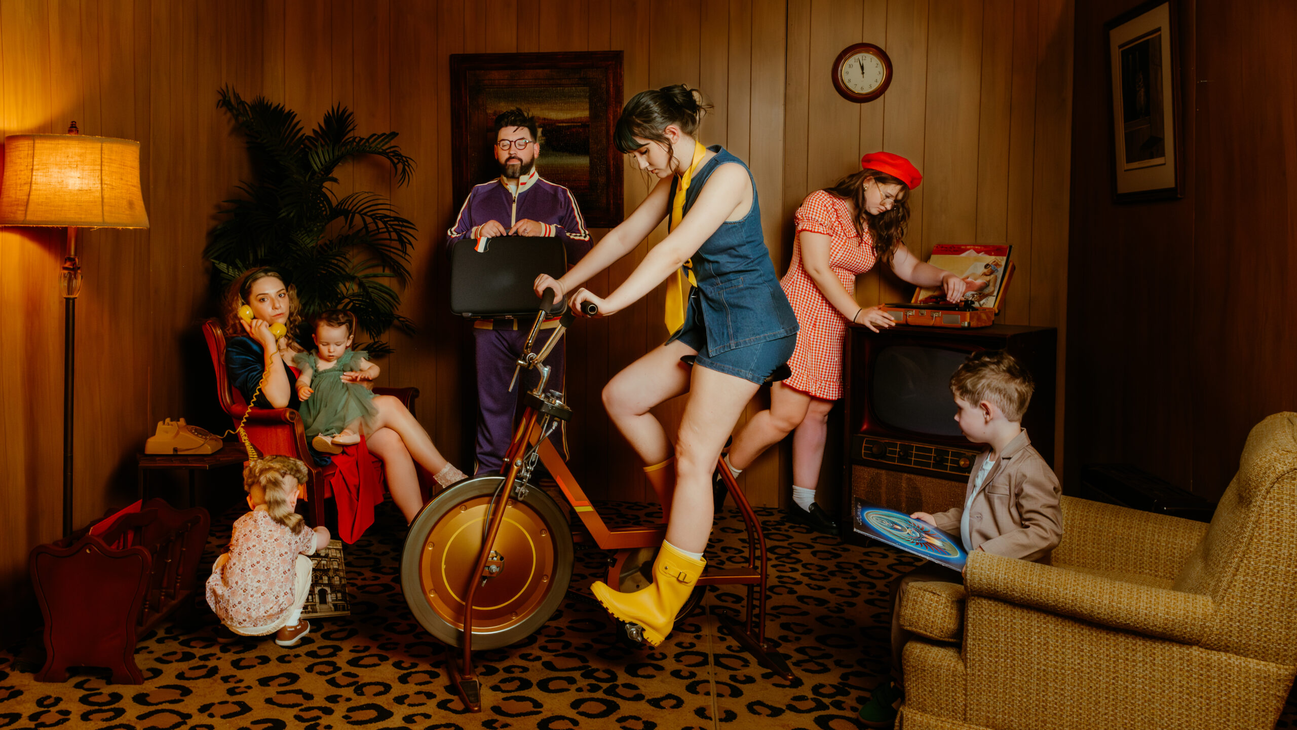 Six people in Wes Anderson-inspired retro clothing gather in a wood-paneled Nashville room with vintage decor. One woman pedals a stationary bike while others relax, read, use a record player, and enjoy the feel of stylish family portraits come to life.