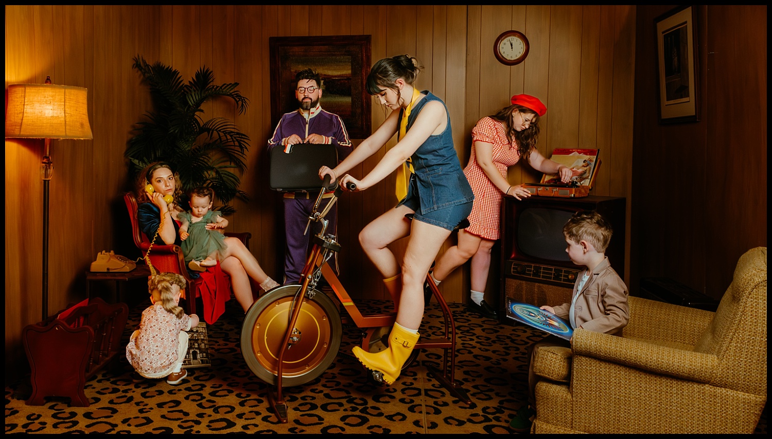 Six people in vintage clothing gather in a warm, wood-paneled room with leopard-print carpet—evoking Wes Anderson’s style. While one woman pedals a stationary bike, others lounge or play as they create quirky family portraits with vinyl records.
