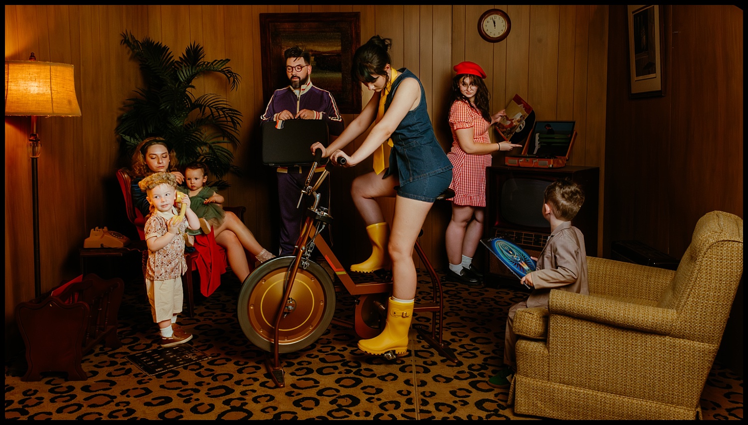 Six people in vintage clothes gather in a retro Nashville living room with wood paneling and leopard-print carpet—a scene reminiscent of Wes Anderson’s family portraits. One woman rides an exercise bike as the others chat, holding records nearby.