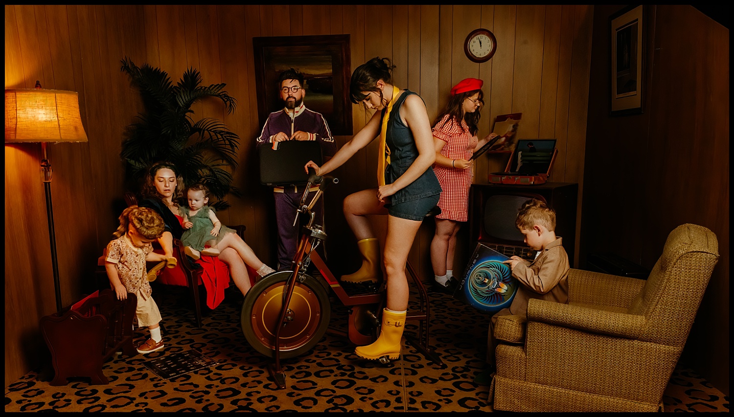 Six people in retro-style clothing pose for Wes Anderson-inspired family portraits in a wood-paneled Nashville room, complete with vintage furniture, a lamp, and potted plant. One woman rides a stationary trike while others sit, read, or play.
