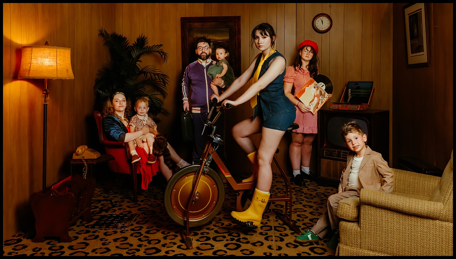 A group of six people, including two children, pose for Nashville-inspired family portraits in a vintage living room with wood panel walls—a scene reminiscent of a Wes Anderson film, complete with retro furniture and quirky yellow boots on a tricycle.