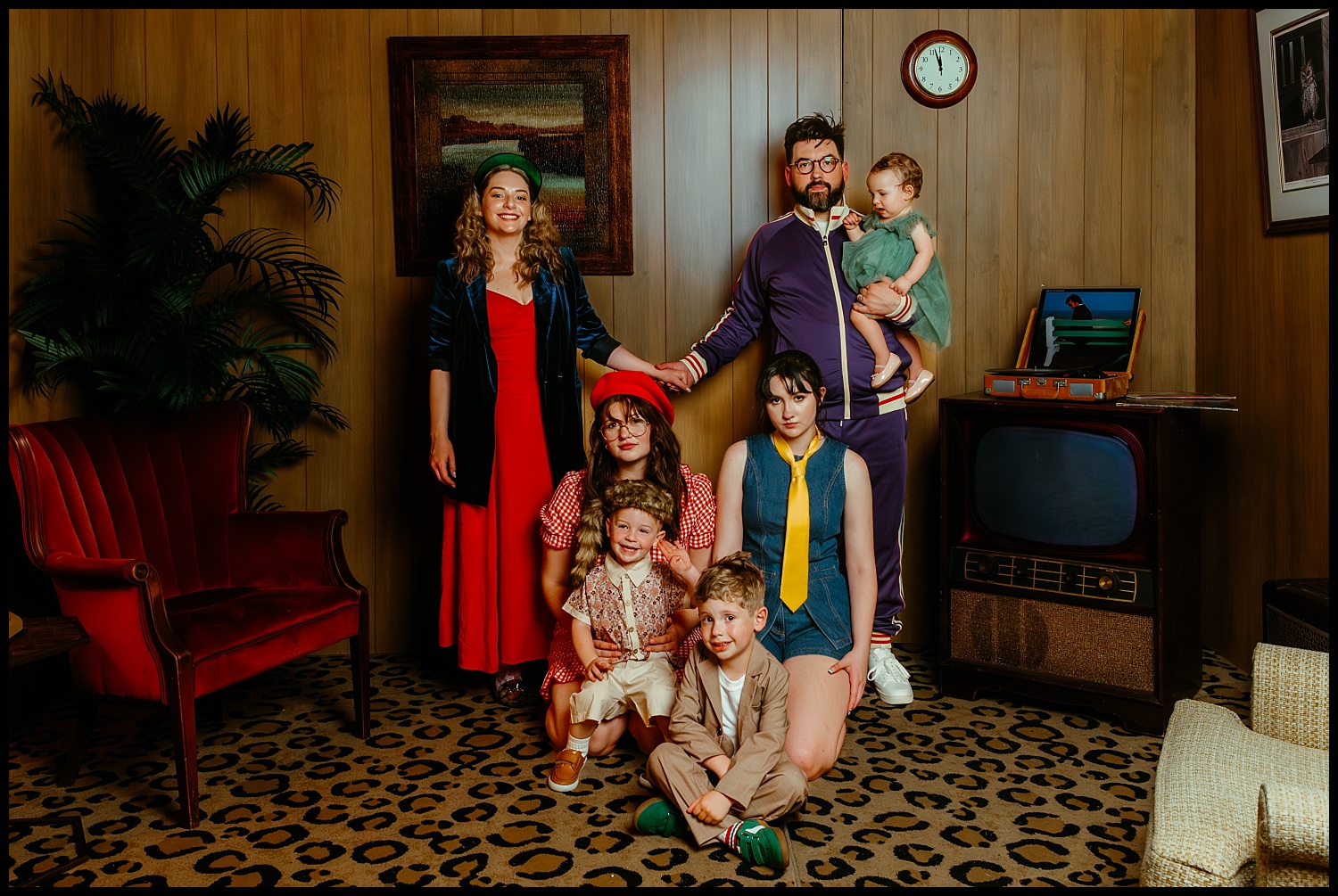 A family of seven poses for quirky Family Portraits in a retro Nashville living room with wood-paneled walls, vintage TV, animal-print carpet, and red velvet chair—channeling a vibrant Wes Anderson aesthetic.