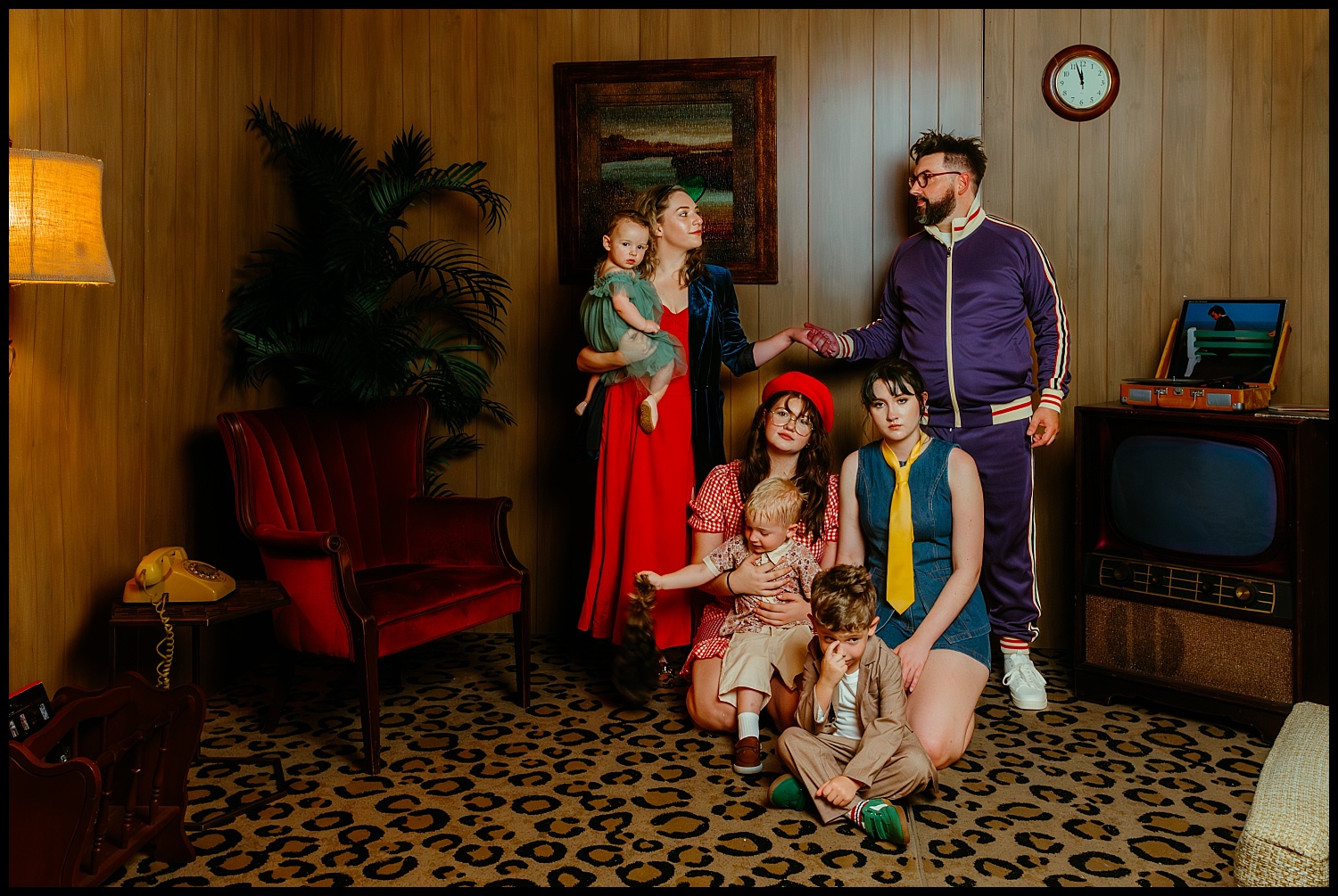 A family of six poses in a retro Nashville living room with wood-paneled walls, vintage furniture, and leopard-print carpet—a Wes Anderson-inspired scene of colorful vintage clothing and quirky family portraits.