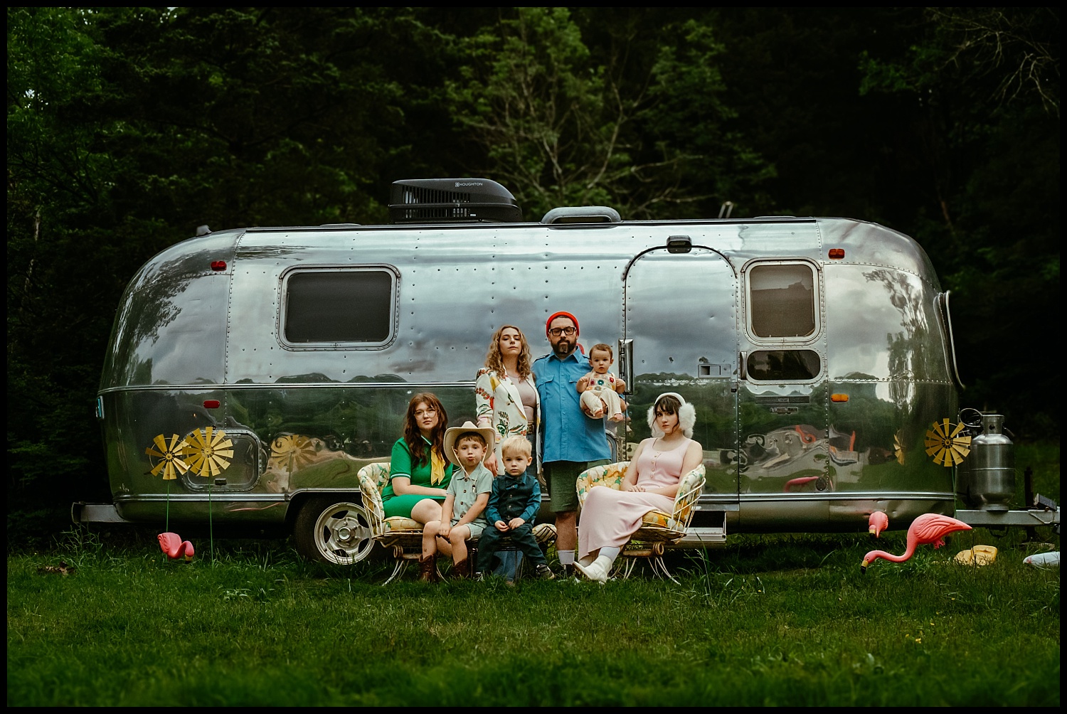 A family of six poses Wes Anderson-style in front of a shiny silver camper trailer surrounded by trees. This whimsical Nashville scene features lawn chairs, toy flamingos, and greenery—perfect for quirky family portraits.