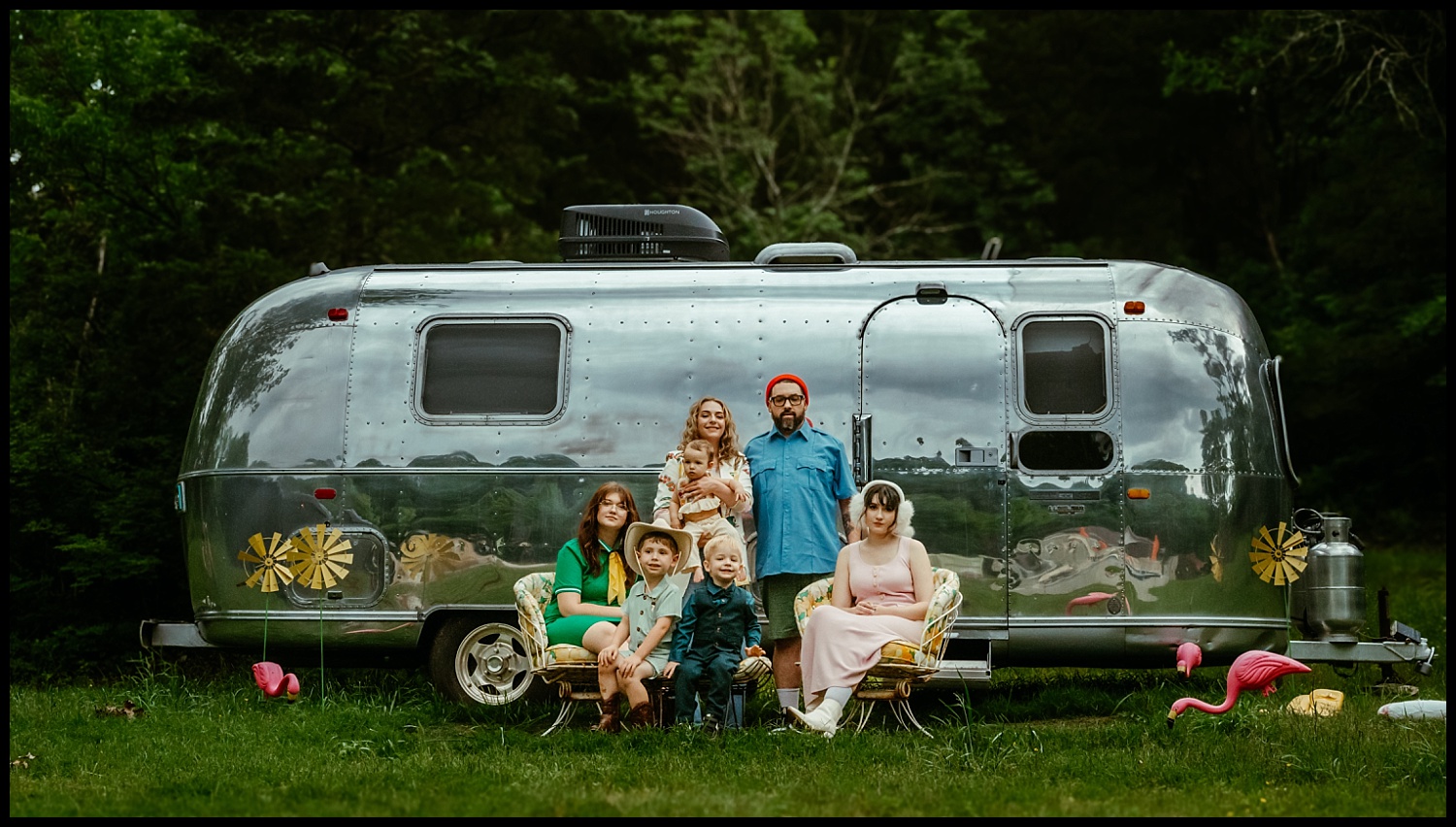 A Wes Anderson-inspired family portrait: six pose in front of a shiny silver camper trailer near Nashville, surrounded by green grass, trees, vintage chairs, and whimsical pink flamingos on the lawn.