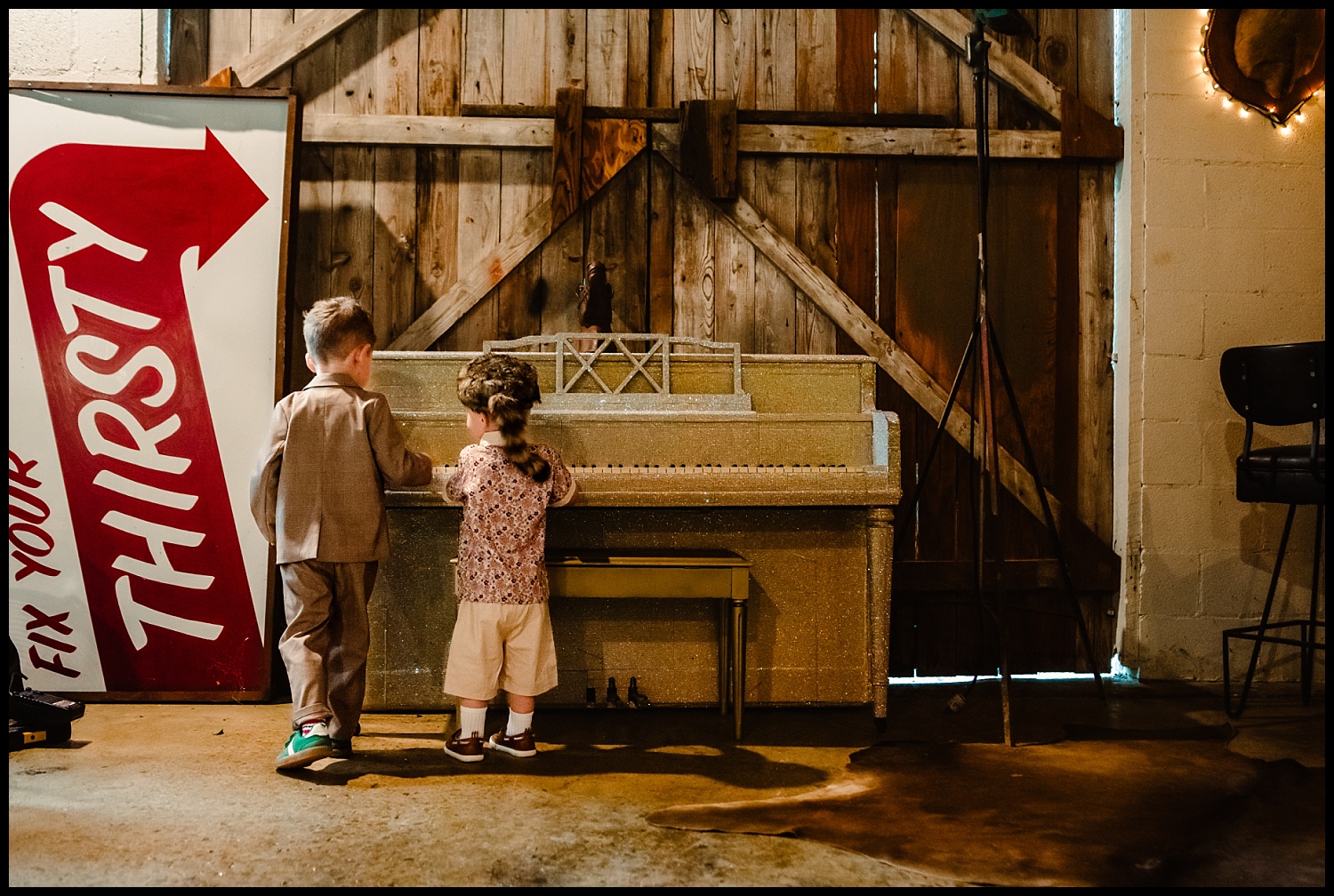 Two young children stand side by side playing a small piano in a rustic room, echoing the quirky charm of a Wes Anderson scene. A large “THIRSTY” sign with a red arrow leans nearby, set against wooden barn doors in the background.