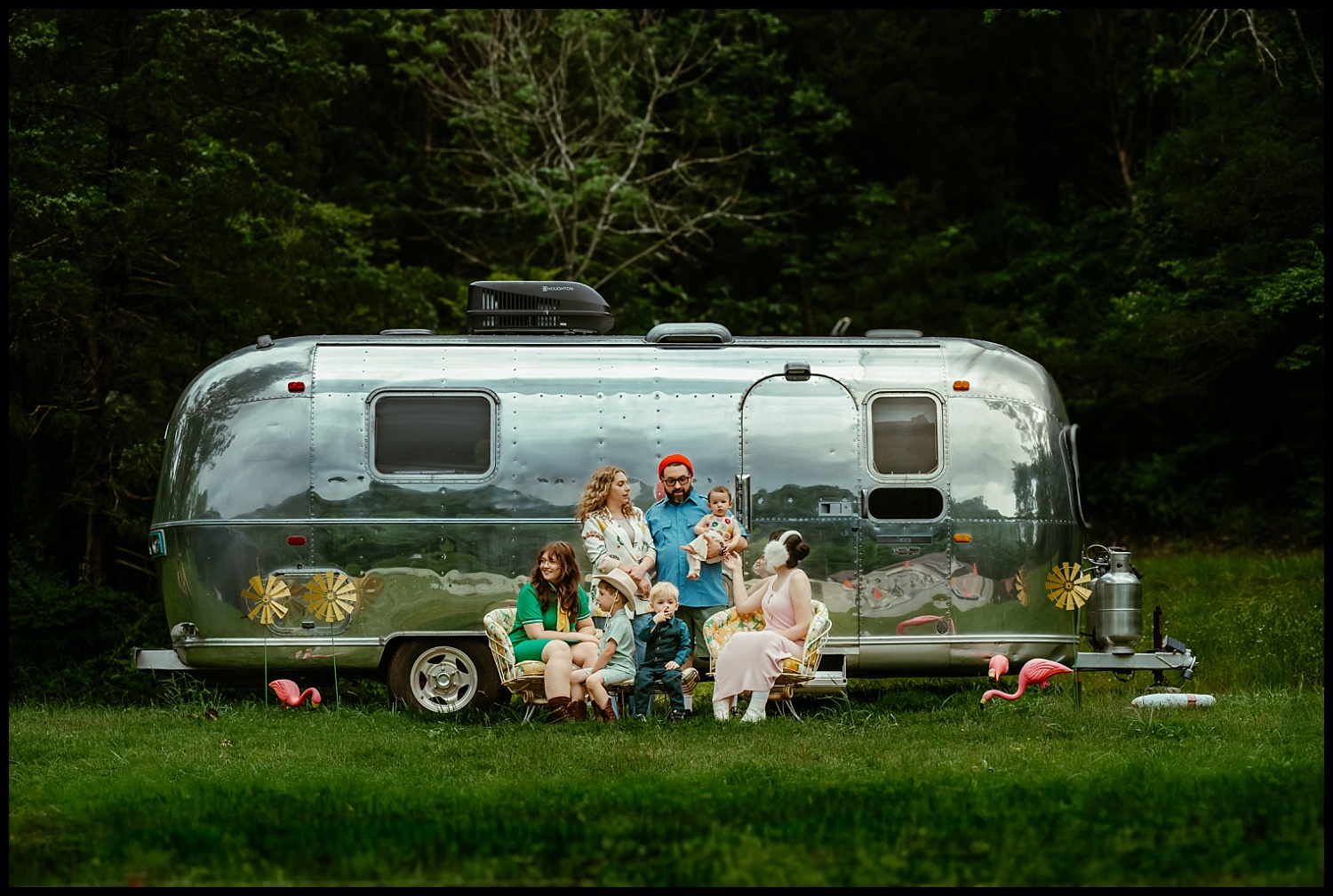 A group of six people, reminiscent of Wes Anderson family portraits, sits and stands together in front of a shiny silver camper trailer in a grassy Nashville area. Two pink flamingo lawn ornaments are visible on the grass.