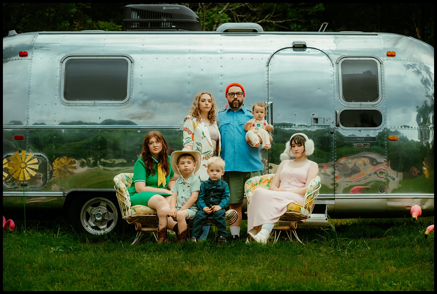 A group of six people, including two children and a baby, pose Wes Anderson-style in front of a shiny silver trailer. Three are seated on patterned chairs, three stand behind, with greenery and trees completing this quirky family portrait.
