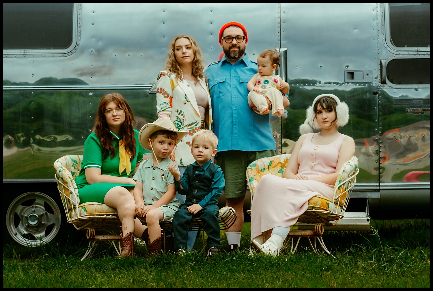 A family of seven poses in front of a shiny silver trailer in true Wes Anderson style. Two adults stand in the back, one holding a baby, while four children sit or stand before them, all dressed in colorful, retro-style clothing.