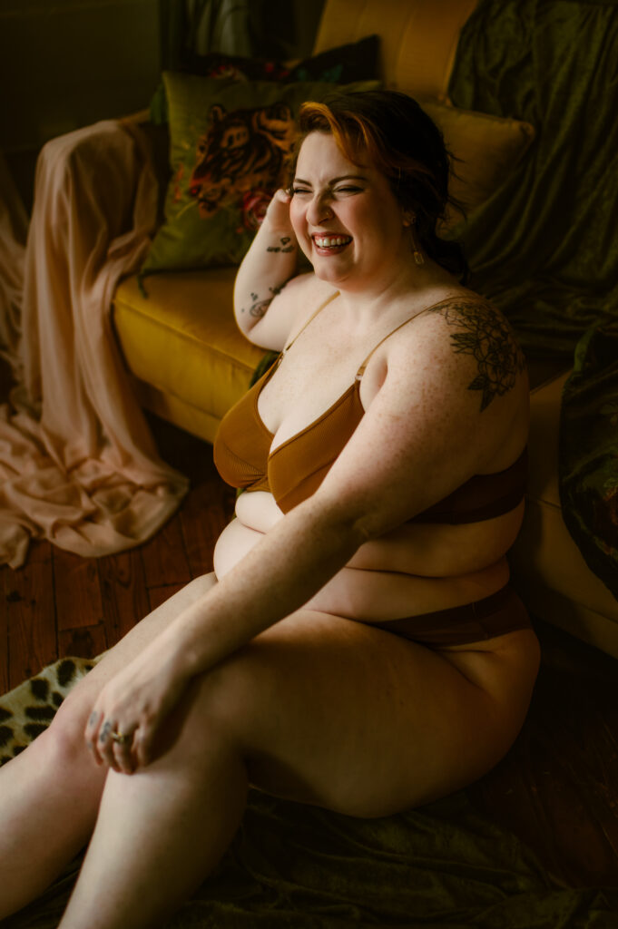 A woman with light skin and short hair sits on the floor in brown lingerie, smiling and looking off to the side. The warm, soft lighting highlights this boudoir photography scene with an expensive yellow couch, green throw, and patterned pillow behind her.