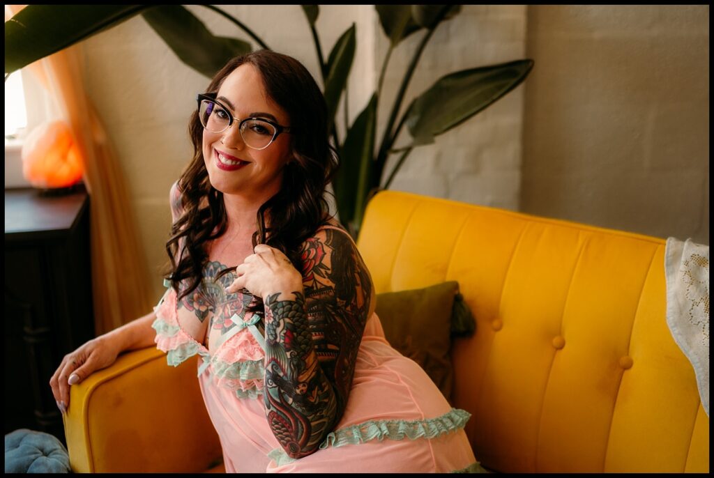 A woman with long dark hair and glasses, wearing a pink and mint green lace robe, smiles while leaning on a bright yellow couch during her Birthday Boudoir Session in a cozy Nashville Studio filled with plants and sunlight.