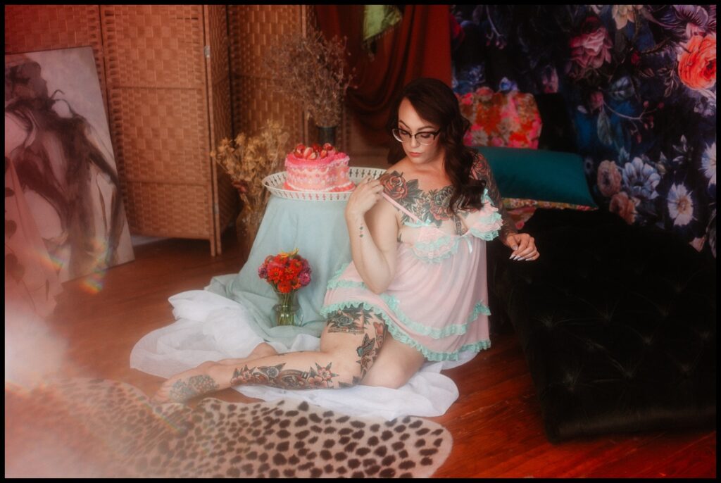 A person with tattoos and glasses poses on the floor in sheer pastel lingerie, surrounded by a floral backdrop, art, flowers, a pink cake on a table, and a leopard print rug during a whimsical Nashville Studio Birthday Boudoir Session.