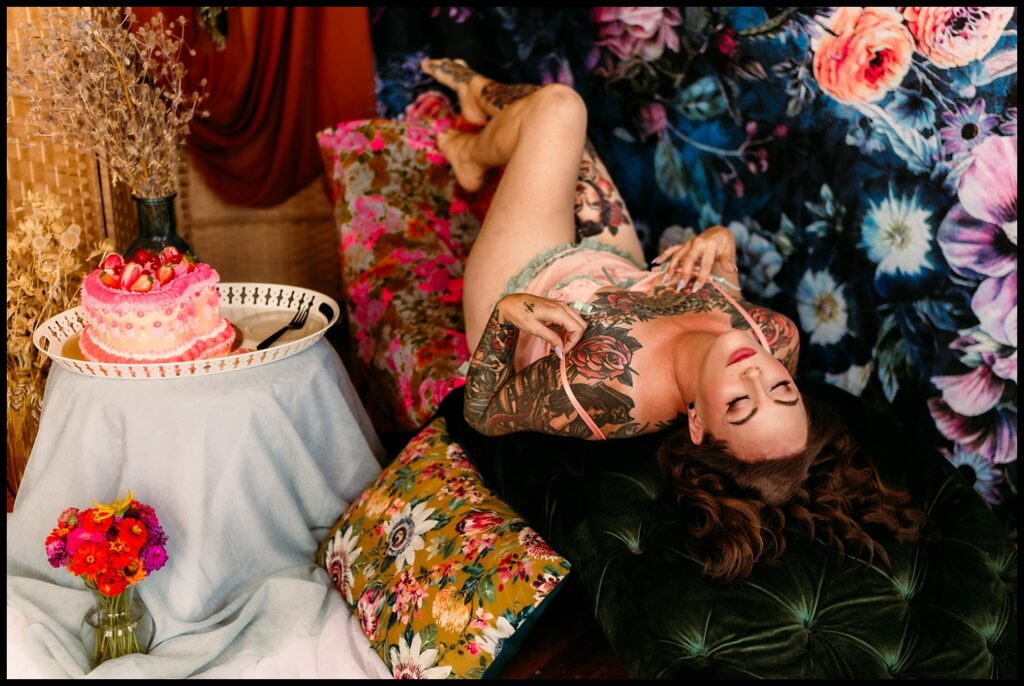 A woman with tattoos reclines upside down on a velvet couch at a Nashville Studio, surrounded by floral decor. A pink decorated cake and a vase of colorful flowers sit nearby, enhancing this vibrant Birthday Boudoir Session scene.