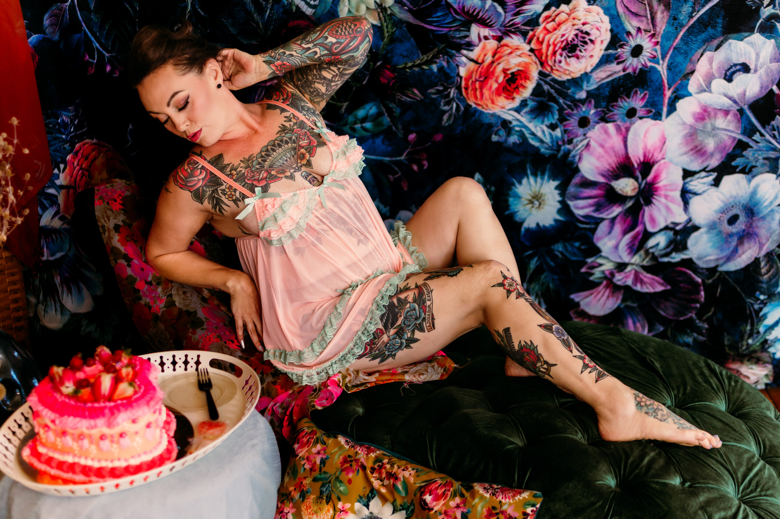 A woman with multiple tattoos lounges on a green ottoman in a pink sheer nightgown, surrounded by vibrant floral decor—a dreamy boudoir session in Nashville, TN. A pink cake with strawberries sits nearby on a white tray, hinting at a special birthday celebration.