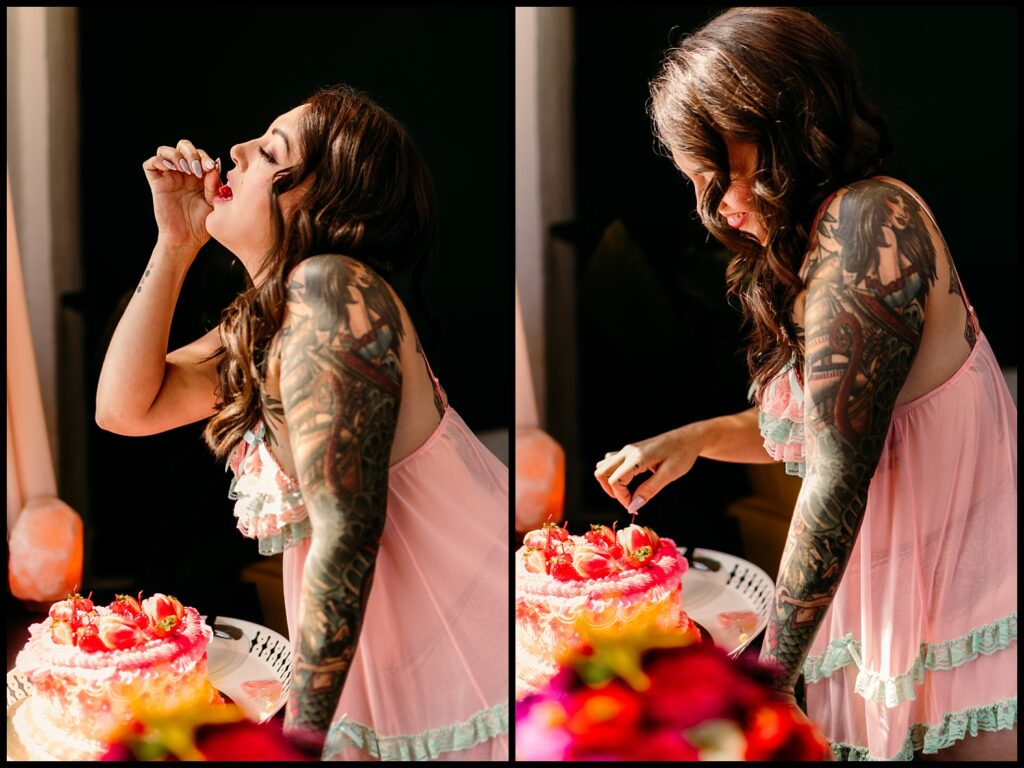 A woman with long hair and colorful tattoos, wearing a pink ruffled dress, tastes frosting from her finger as she decorates a pink frosted cake with strawberries during a Birthday Boudoir Session in a sunlit Nashville Studio.