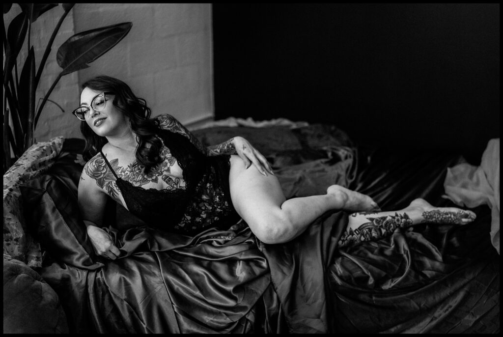 A woman with glasses and tattoos poses confidently on a bed covered with silky sheets in a Nashville Studio, wearing a lace bodysuit. The black-and-white photo captures the relaxed, intimate vibe of a Birthday Boudoir Session.
