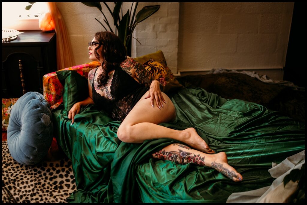 A woman with long dark hair and tattoos lounges on a green satin bedspread in a Nashville Studio, wearing black lace lingerie and glasses. She looks off to the side in a softly lit, cozy room with colorful pillows and decor.