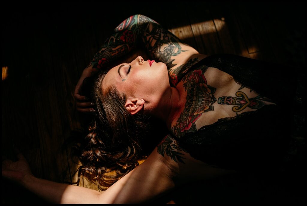 A woman with tattoos lies on a wooden floor in sunlight and shadow at a Nashville Studio. She is wearing a black lace top, eyes closed, with one arm extended and her hair spread out—perfect for a Birthday Boudoir Session.