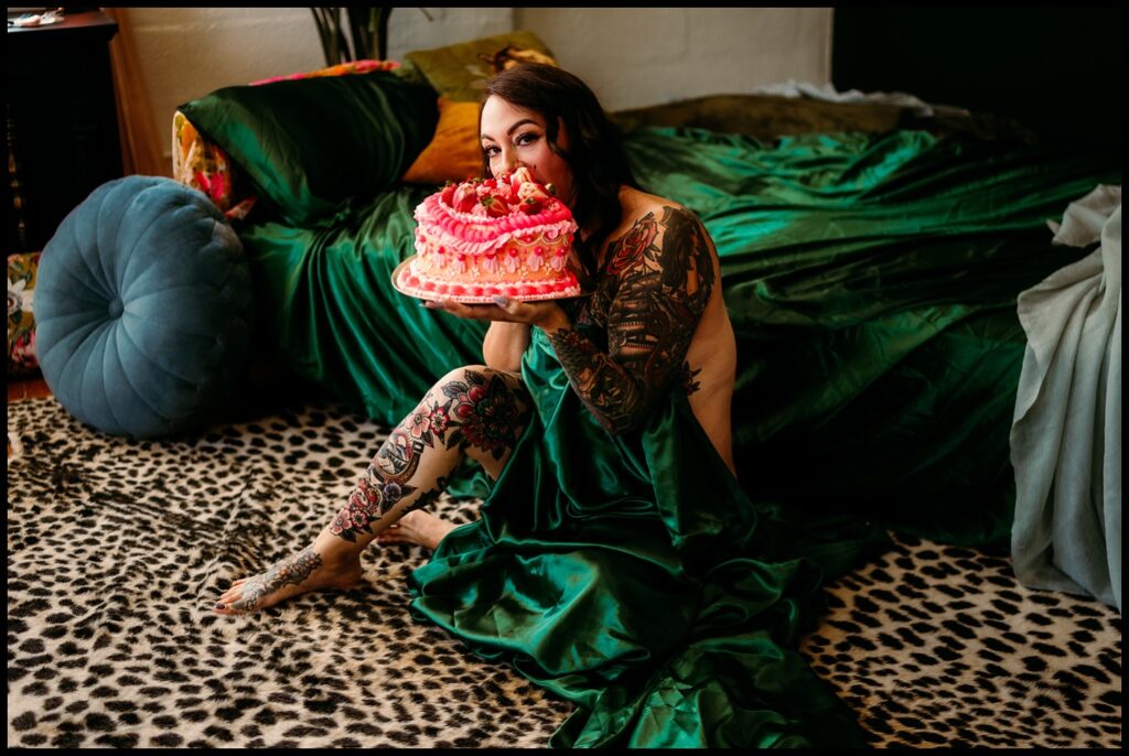 A tattooed woman sits on a cheetah-print rug at a Nashville Studio, partially wrapped in a green satin sheet, holding a pink decorative cake and smiling, with a green bed and colorful pillows behind her—a perfect Birthday Boudoir Session.