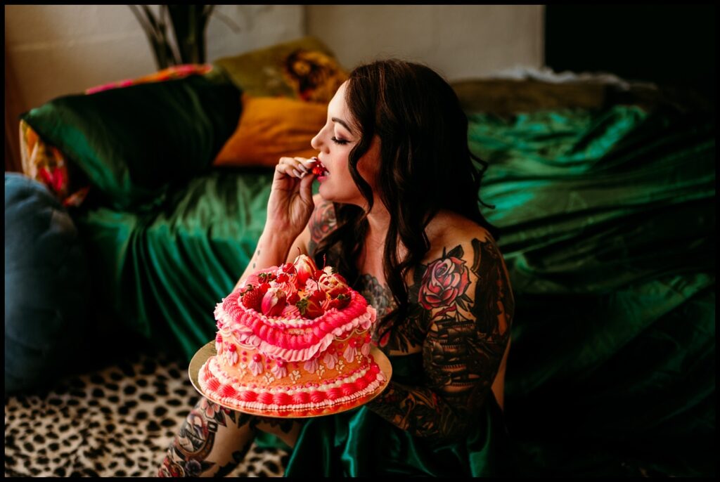A woman with long dark hair and colorful tattoos sits on a bed with green sheets in a Nashville studio, holding a pink and yellow decorated cake and tasting some with her fingers—a playful birthday boudoir session.