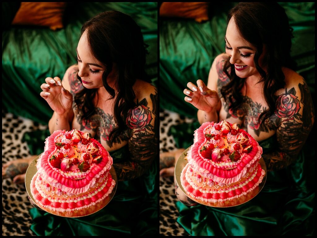 A woman with long dark hair and colorful tattoos smiles while holding a pink, heart-shaped cake topped with strawberries during her Birthday Boudoir Session at a Nashville Studio, sitting on a green surface with a black-and-white rug beneath her.