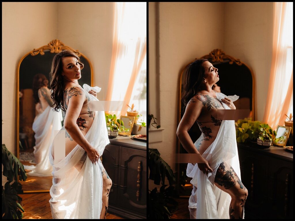 A woman draped in a white sheet poses confidently during a Birthday Boudoir Session in a sunlit Nashville studio, with plants and a large ornate mirror revealing her tattoos and reflecting her image.