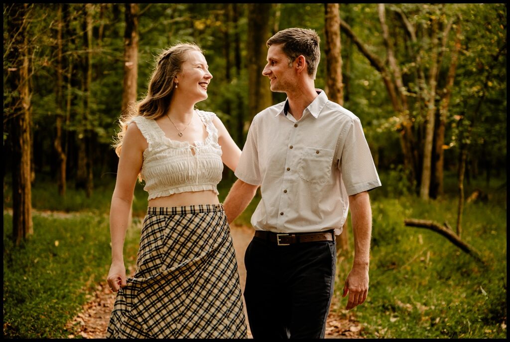 A smiling couple walks hand-in-hand down a wooded path during golden hour. The woman wears a white crop top and plaid skirt, the man a white shirt and dark pants. They share a romantic gaze, surrounded by trees and lush greenery.