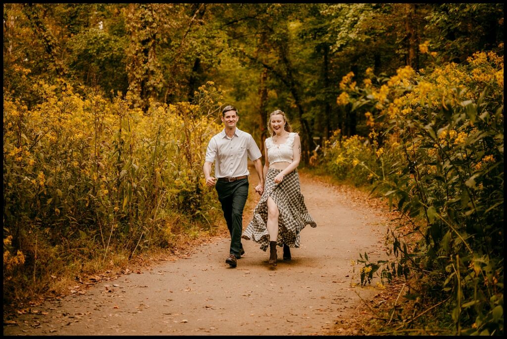 A man and woman walk together down a path surrounded by tall yellow wildflowers and green trees, both smiling and enjoying a sunny golden hour during their romantic session outdoors.
