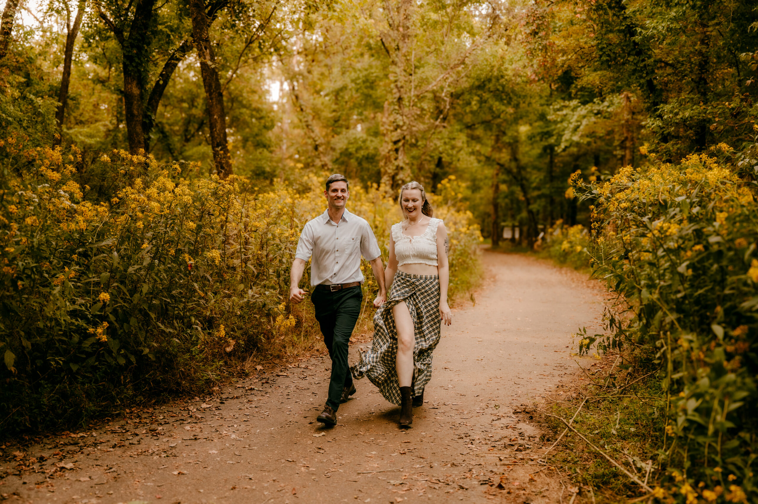 A couple walks hand in hand down a tree-lined path surrounded by yellow wildflowers during golden hour. Both are smiling; the woman wears a flowy skirt and boots, and the man wears a white shirt and dark pants in this warm, inviting romantic session.