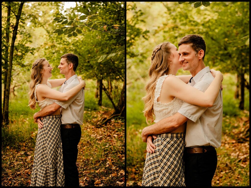 A couple stands in a sunlit forest during golden hour, embracing and smiling at each other. The woman wears a plaid skirt and white top, while the man wears a light shirt and dark pants. They look happy and affectionate in the greenery.