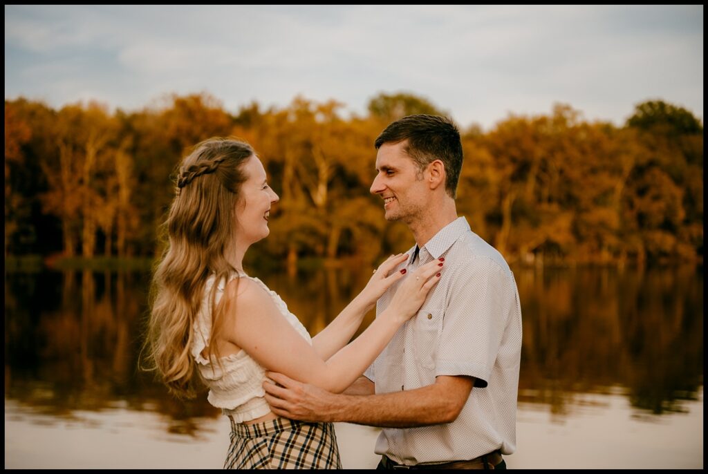 A couple stands close together, smiling at each other by a calm lake during golden hour, with autumn trees and warm sunlight setting the scene for their romantic session.