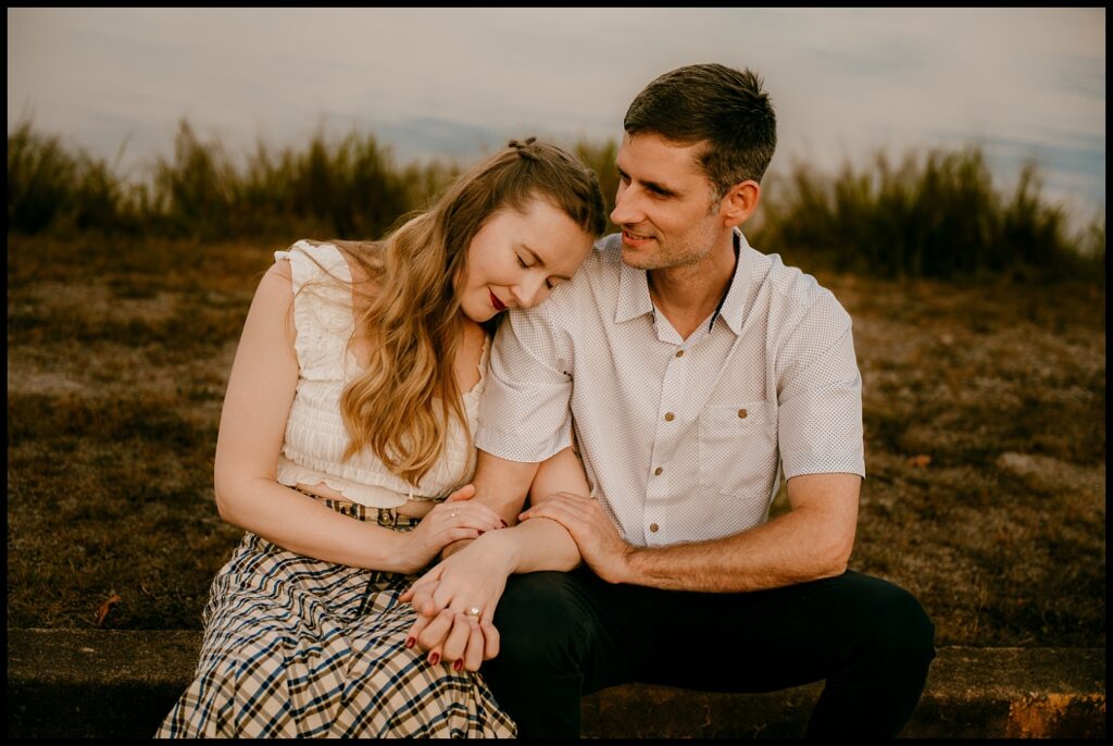 A woman rests her head on a man's shoulder as they sit together outdoors during golden hour, holding hands and smiling gently. The background shows grass and a cloudy sky, creating a warm, peaceful atmosphere.