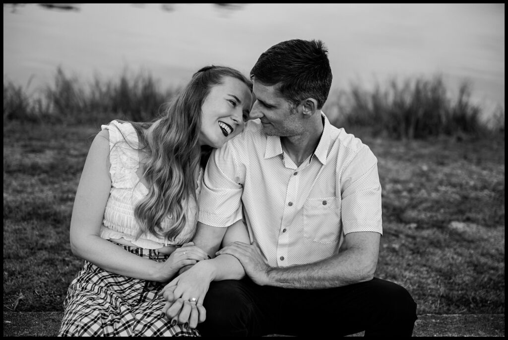 A woman and a man sit close together outdoors during their Nashville engagement session, smiling and holding hands. They gaze at each other affectionately, with grass and water softly blurred in the black-and-white background.