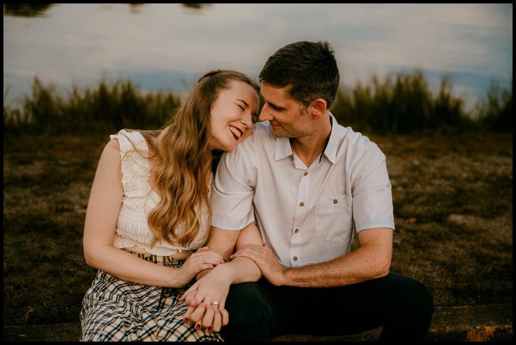 A couple sits close together outdoors during golden hour, holding hands and smiling warmly at each other. The woman leans affectionately on the man's shoulder as grass and water blur softly in the background of their romantic Nashville engagement session.