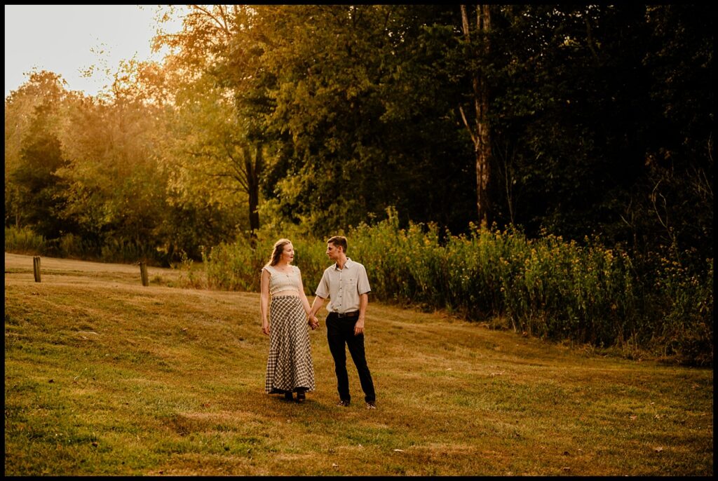 A couple holding hands walks through a grassy field at golden hour, surrounded by trees and tall grass near Nashville, with warm light illuminating their engagement session.