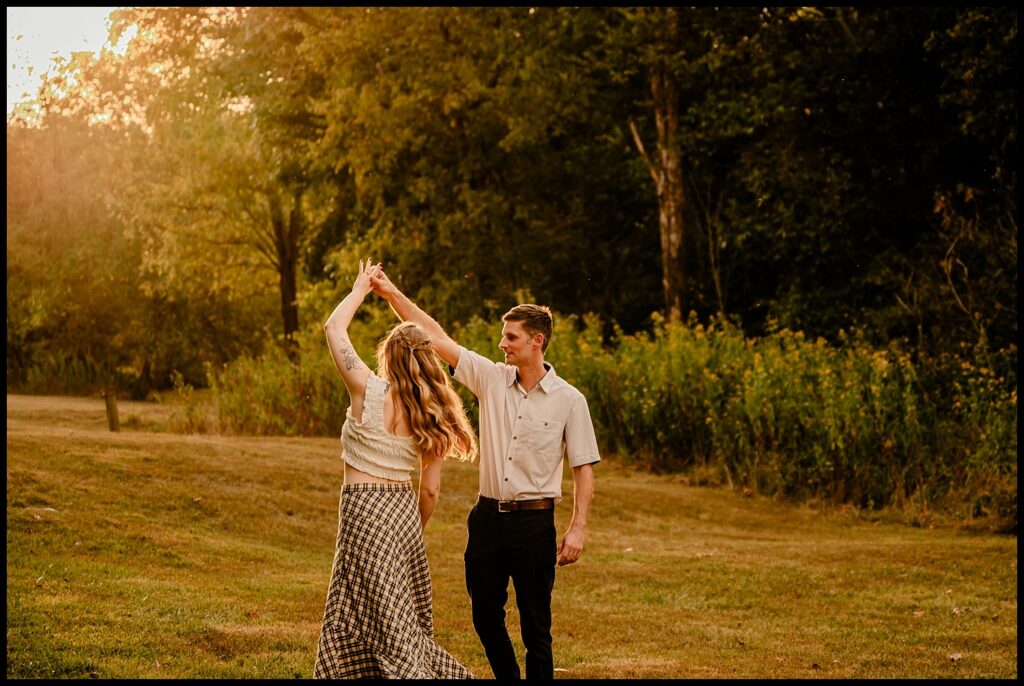 A couple dances together outdoors on a grassy field at golden hour, surrounded by trees. The woman twirls with her arm raised, while the man holds her hand and smiles—capturing a romantic session in the warm, golden glow.