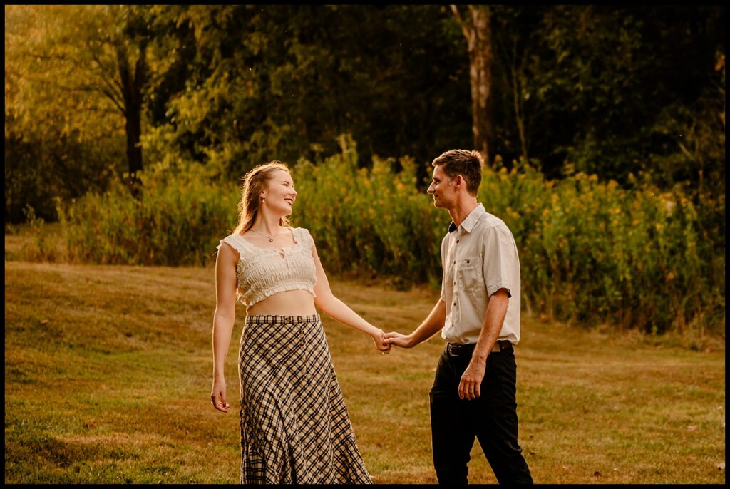 A couple stands outdoors on grass, holding hands and smiling at each other during their Nashville engagement session. They are surrounded by trees and greenery under warm, golden hour sunlight.