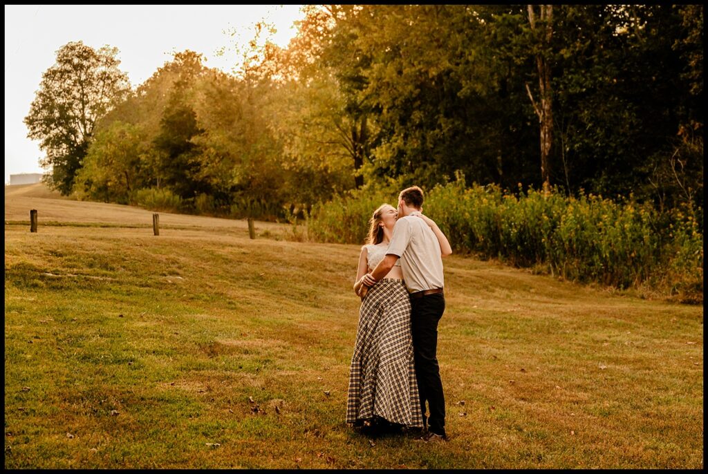 A couple stands in a grassy field at sunset during a romantic session, embracing and sharing a kiss. The woman wears a plaid skirt and white top; the man wears dark pants and a light shirt. Trees and golden hour sunlight fill the background.