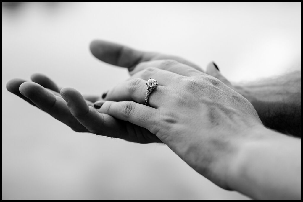 A close-up, black-and-white photo of two hands gently touching, with one hand wearing a diamond engagement ring. The blurred background draws focus to the hands and ring, capturing the intimacy of a Nashville engagement.