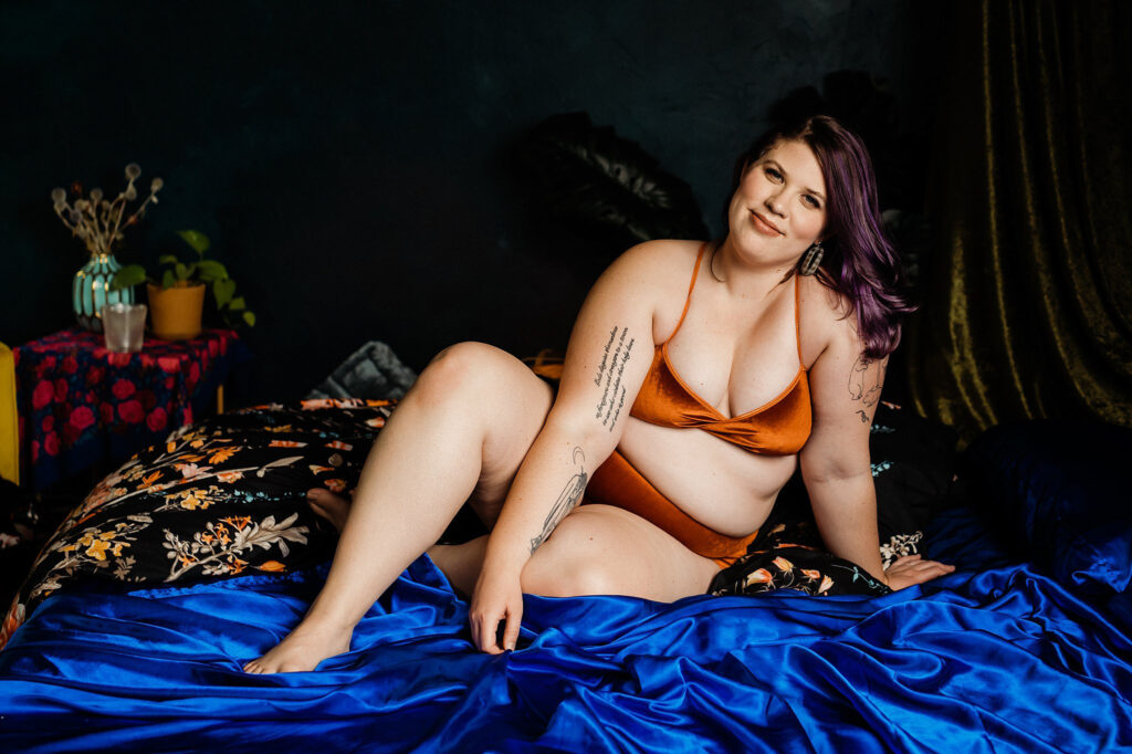 A woman with purple hair wearing a copper-colored bikini sits confidently on a bed with vibrant blue and floral bedding, surrounded by dark, moody decor and plants. She is smiling, her tattoos visible—a true Nashville boudoir vibe.