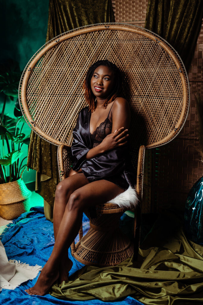 A woman in a black lace lingerie set and silky robe sits on a large wicker chair, surrounded by green curtains and indoor plants, with colorful fabrics on the floor. She is smiling and looking to the side in this Nashville boudoir scene.