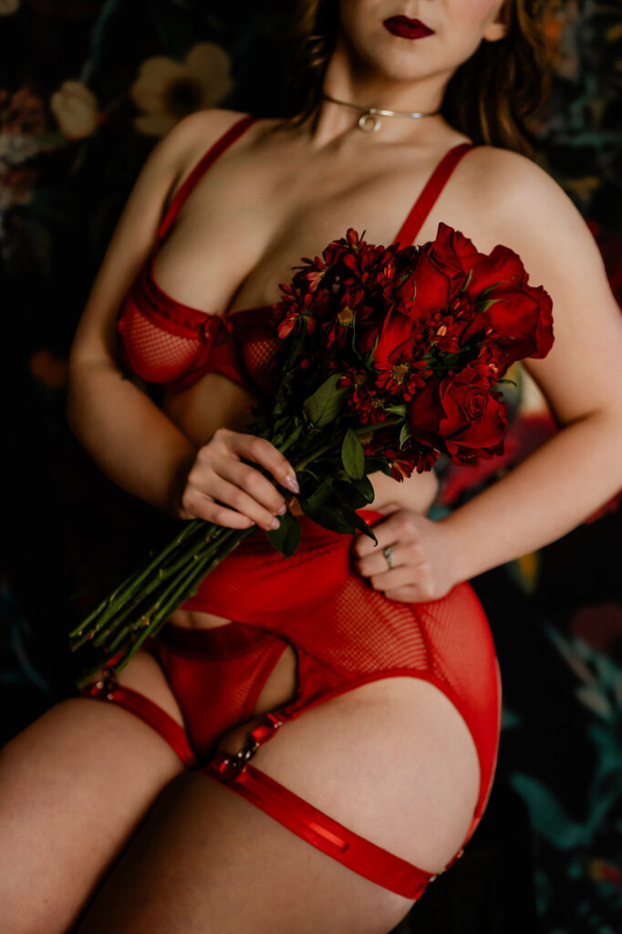 A woman in red lingerie and a choker necklace holds a bouquet of red roses against a dark, floral background. Her face is partially visible, capturing the intimate, moody vibe of a Nashville boudoir setting.
