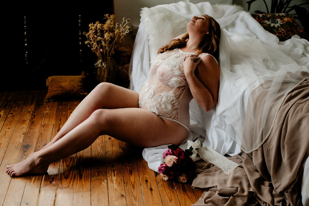 A woman in a sheer lace bodysuit sits on a wooden floor, leaning back against a bed draped with white and beige fabric, holding her chest and looking upward—a soft, elegant moment from a Nashville boudoir session. A bouquet of flowers rests beside her.