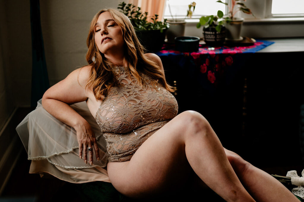 A woman with long, light brown hair poses on a chair in a beige lace bodysuit for a Nashville boudoir session. She sits with her eyes closed, one arm resting on the chair, as light streams in from a window lined with plants behind her.