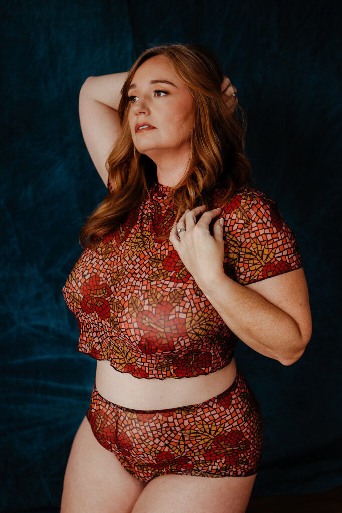 A woman with long auburn hair poses confidently in a matching red and gold mosaic-patterned sheer crop top and high-waisted underwear, capturing the essence of a Nashville boudoir session against a dark blue textured background.